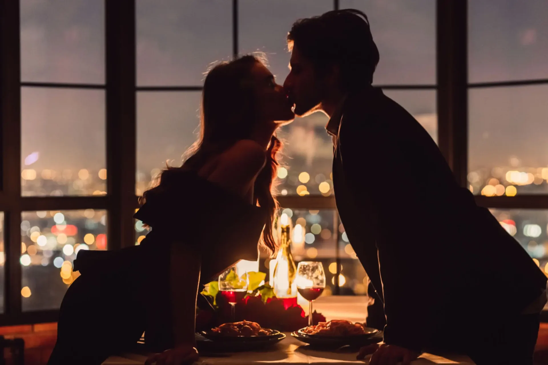 A romantic couple sharing a kiss at a candlelit dinner with city lights in the background.